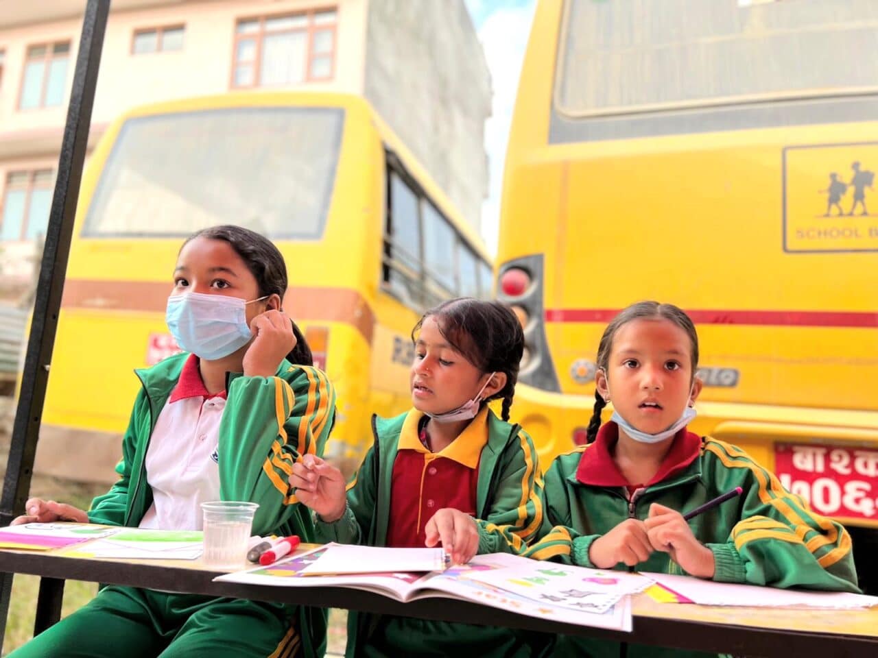 image of 3 young girls sitting at a table drawing. yellow buses behind them