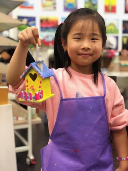 image of student holding a painted wooden house with purple apron